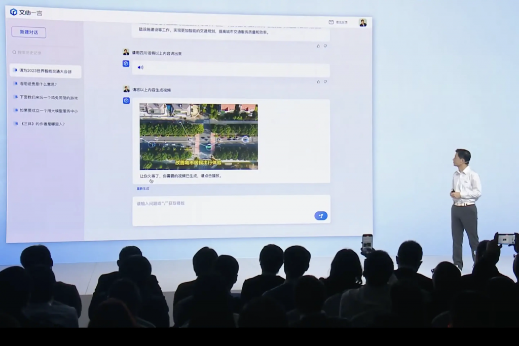Ein Mann präsentiert auf einer Technologiekonferenz neben einem großen Bildschirm, der eine Social-Media-Oberfläche zeigt, neue KI-Modelle während das Publikum Fotos macht. Der Bildschirm zeigt chinesischen Text, was auf einen Schwerpunkt auf digitale Technologien hinweist.
