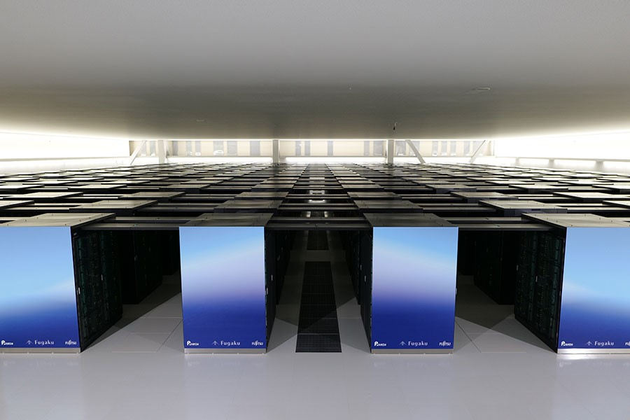 A modern interior view of the Fugaku supercomputer shows a symmetrical arrangement of reflective, upright panels labeled 'Fugaku'. The ceiling is evenly lit, highlighting the geometric precision of the layout in this state-of-the-art facility.
