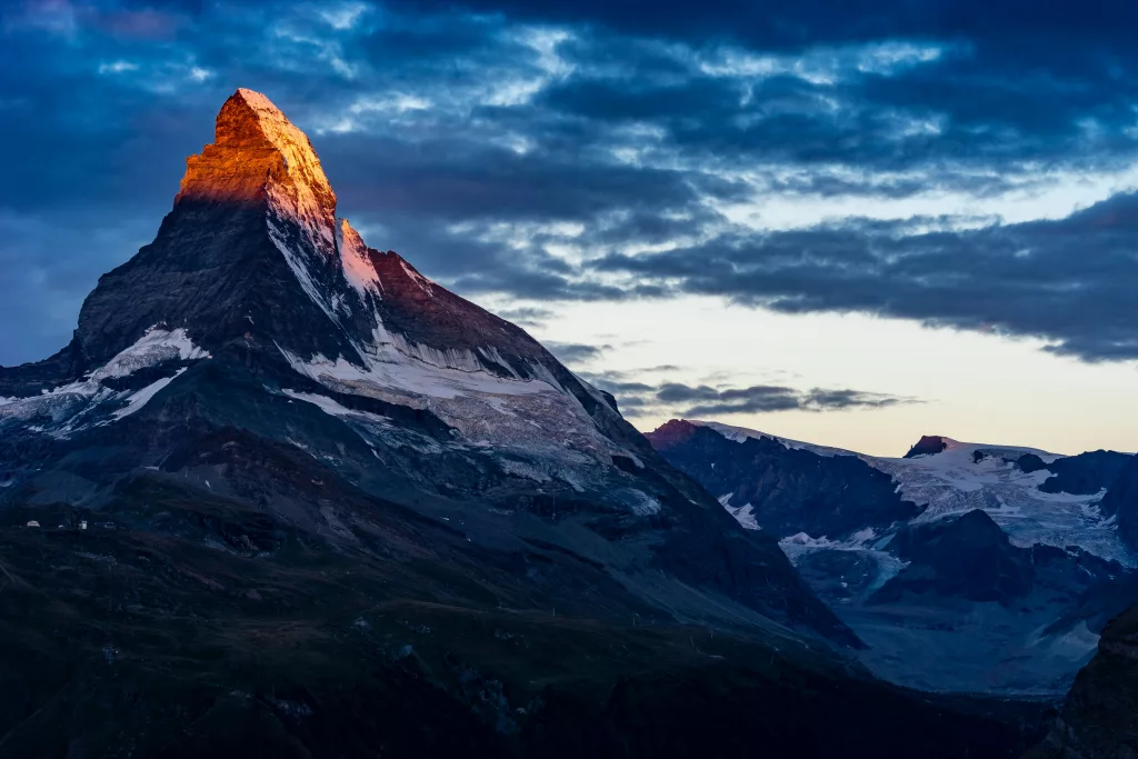 Matterhorn, einen der bekanntesten Berge der Alpen, der sich an der Grenze zwischen der Schweiz und Italien befindet. Der Gipfel des Matterhorns ist in warmes Sonnenlicht getaucht, das ihm eine goldene und orangefarbene Färbung verleiht. Der Rest des Berges und der umliegenden Landschaft ist im Schatten, mit dramatischen Wolken, die den Himmel bedecken und dem Bild eine beeindruckende Atmosphäre verleihen. Der Schnee und das Eis auf dem Matterhorn und den umliegenden Gipfeln sind deutlich sichtbar, was die majestätische und unberührte Schönheit dieser Berglandschaft unterstreicht.