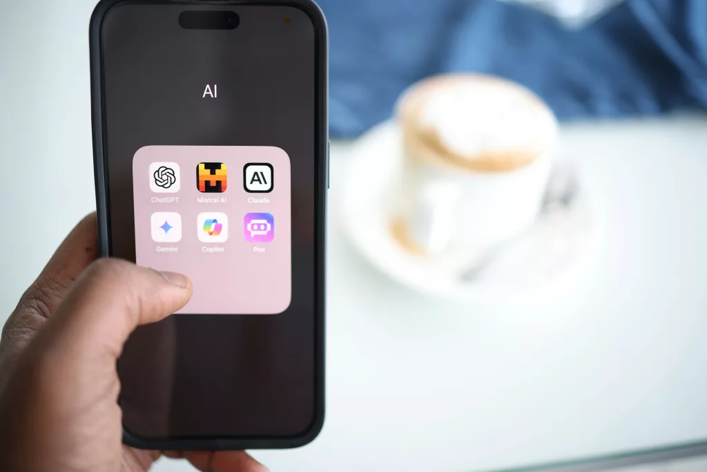 Ein Smartphone wird von einer Hand gehalten und zeigt einen Bildschirm mit einem Ordner namens "AI", der die Apps ChatGPT, Mistral AI, Claude, Gemini, Copilot und Poe enthält. Im Hintergrund ist eine unscharfe Tasse Kaffee auf einem Unterteller und ein blaues Tuch zu sehen.